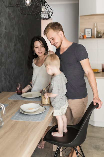 Smiling young couple looking at little boy standing on chair serving oats on plate at breakfast