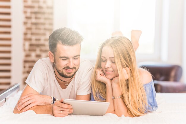 ベッドに横たわっている笑顔の若いカップルは、ベッドルームのデジタルタブレットを見て
