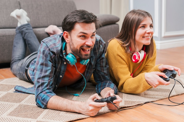 Улыбающаяся молодая пара лежит на полу и играет в видеоигру с джойстиком у себя дома