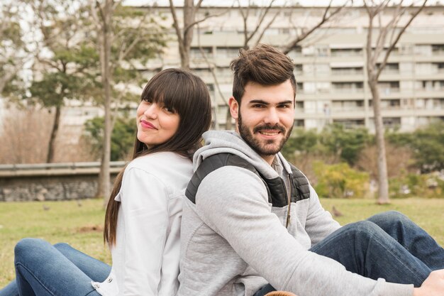 Улыбается молодая пара, сидя спиной к спине в парке