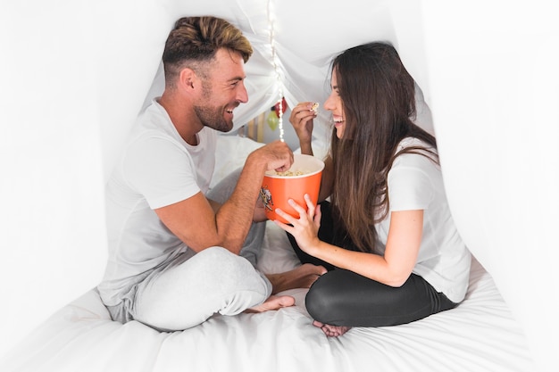 Smiling young couple sitting on bed covered with white curtain eating popcorn