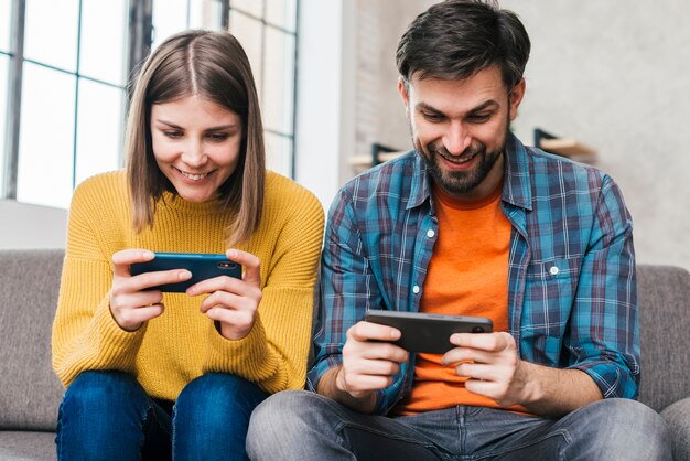 携帯電話でビデオゲームをプレイソファーに座っていた若いカップルの笑顔 プレミアム写真 携帯電話でビデオゲームをプレイソファーに座っていた若いカップルの笑顔 プレミアム写真