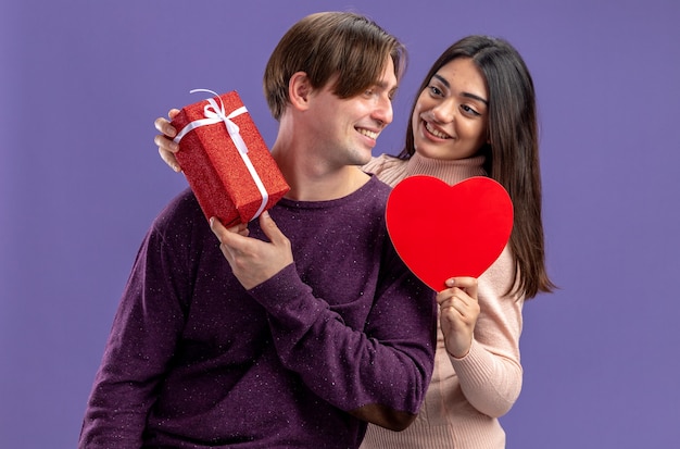 Smiling young couple on valentines day looking at each other holding heart shaped box with gift box isolated on blue background