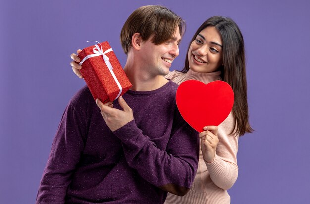 발렌타인 데이에 웃는 젊은 부부는 파란색 배경에 격리된 선물 상자가 있는 하트 모양의 상자를 들고 서로를 바라보고 있습니다.