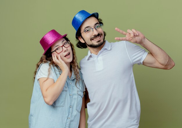 Smiling young couple wearing pink and blue hat girl putting hand on cheek and guy showing peace gesture isolated on olive green