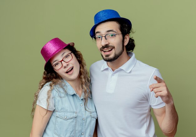 Smiling young couple wearing pink and blue hat guy showing you gesture isolated on olive green