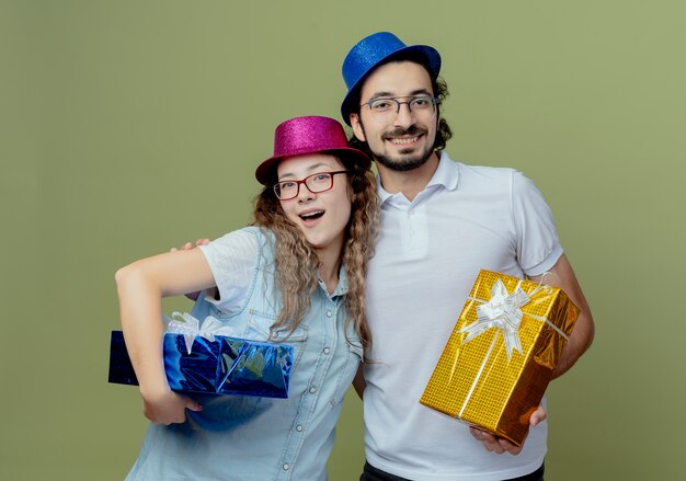 Smiling young couple wearing pink and blue hat hug each other and holding gift boxes isolated on olive green wall