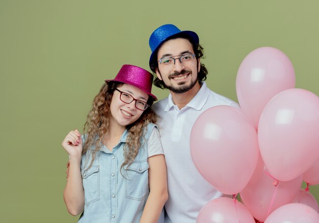 Smiling young couple wearing pink and blue hat standing nearby balloons isolated on olive green