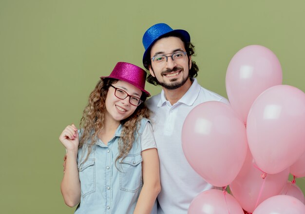 Улыбающаяся молодая пара в розово-голубой шляпе стоит рядом с воздушными шарами, изолированными на оливково-зеленом