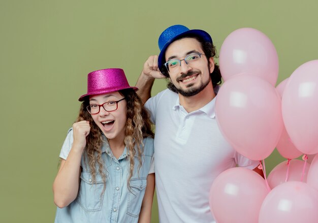 Smiling young couple wearing pink and blue hat standing nearby balloons and showing yes gesture isolated on olive green