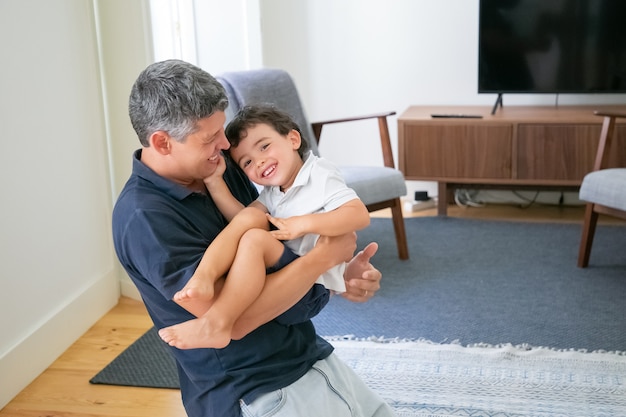 Улыбающийся молодой папа держит сына на руках и стоит на коленях в гостиной.