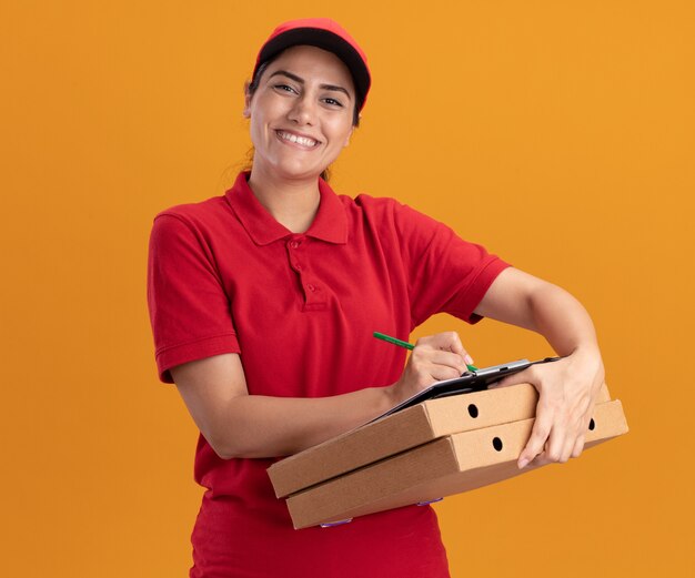 オレンジ色の壁に分離されたピザの箱のクリップボードに何かを書いている制服とキャップを身に着けている正面を見て笑顔の若い配達の女の子