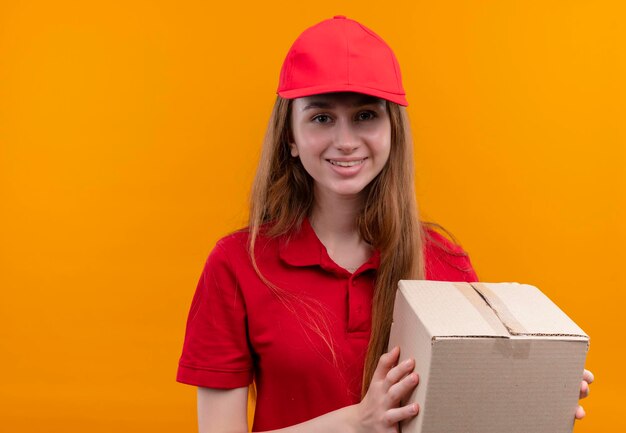 Улыбающаяся молодая доставщица в красной форме, держащая коробку на изолированном оранжевом пространстве с копией пространства
