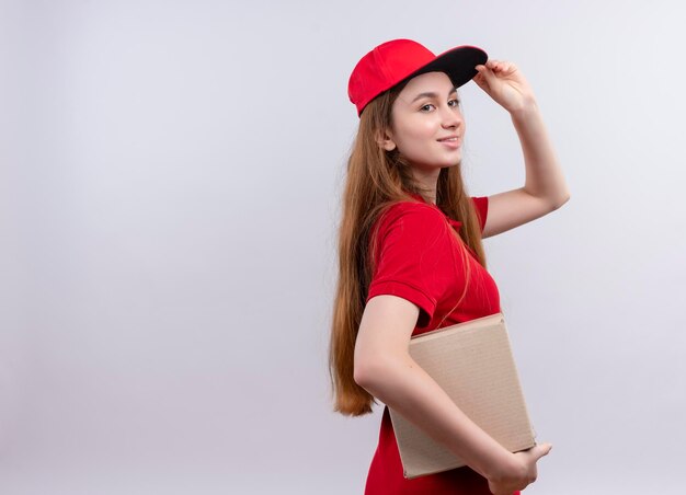 Smiling young delivery girl in red uniform holding box and putting hand on cap standing in profile view on isolated white space with copy space