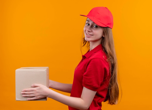 격리 된 주황색 공간에 프로필보기에 서있는 빨간색 유니폼 들고 상자에 젊은 배달 소녀 미소