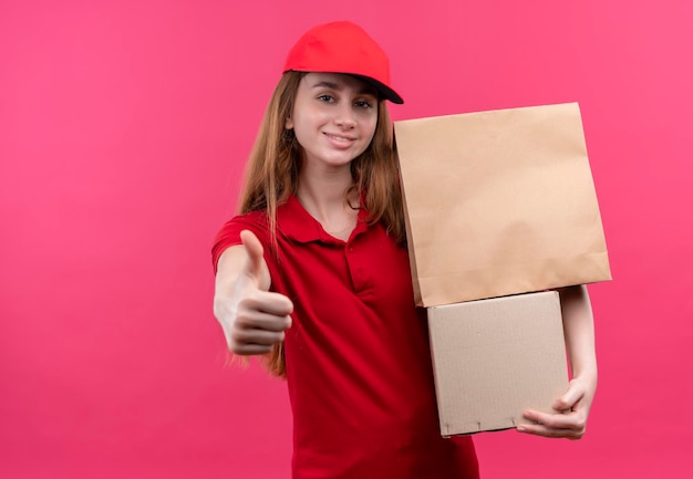 Smiling young delivery girl in red uniform holding boxes and showing thumb up on isolated pink space with copy space