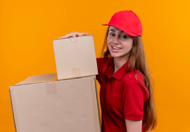 Foto gratuita sorridente giovane ragazza di consegna in rosso uniforme tenendo le scatole in piedi in vista di profilo su uno spazio arancione isolato