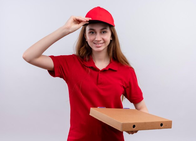 Smiling young delivery girl in red uniform holding package and putting hand on cap on isolated white space