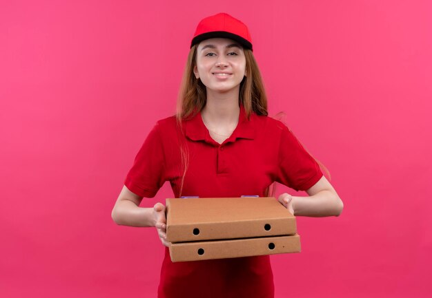 Smiling young delivery girl in red uniform holding packages on isolated pink space with copy space