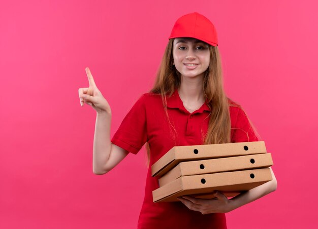 Улыбающаяся молодая доставщица в красной форме держит пакеты и указывает вверх на изолированное розовое пространство с копией пространства