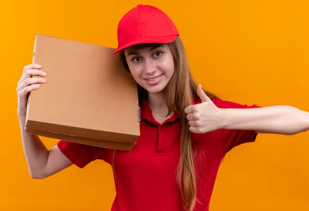 Foto gratuita sorridente giovane ragazza di consegna in uniforme rossa che tiene i pacchetti sulla spalla e che mostra il pollice in su sullo spazio arancione isolato