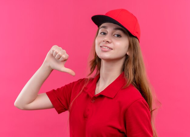 Smiling young delivery girl in red uniform pointing at herself on isolated pink wall