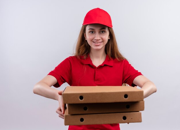 Smiling young delivery girl in red uniform stretching out packages  on isolated white wall