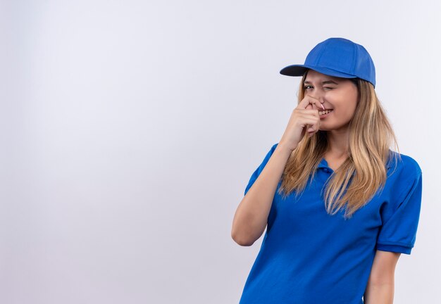 Улыбающаяся молодая доставщица в синей форме и кепке с закрытым носом изолирована на белой стене с копией пространства
