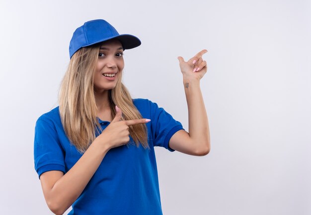 Улыбающаяся молодая доставщица в синей форме и кепке указывает в сторону, изолированную на белой стене с копией пространства