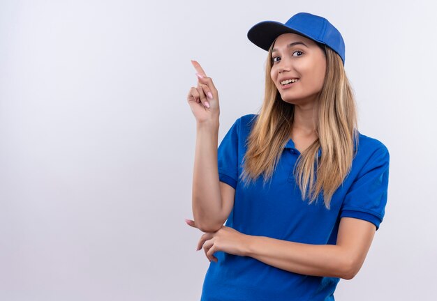 Smiling young delivery girl wearing blue uniform and cap points to side isolated on white wall with copy space