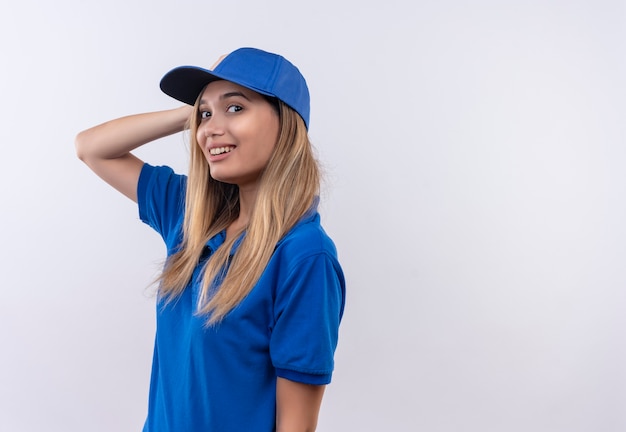 Smiling young delivery girl wearing blue uniform and cap putting hand on head isolated on white