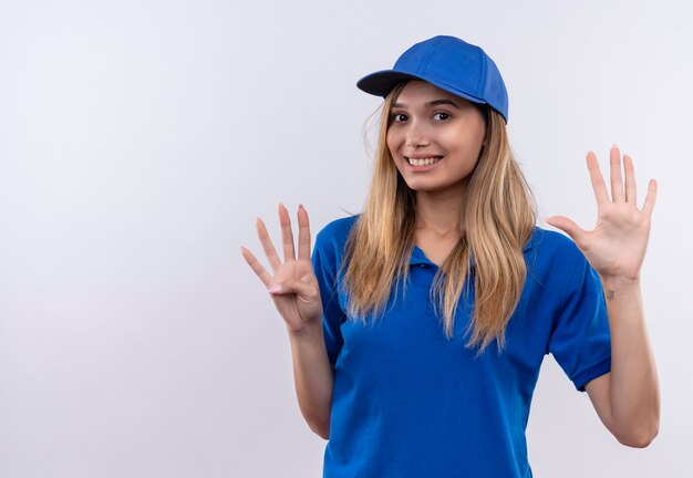 Smiling young delivery girl wearing blue uniform and cap showing different numbers isolated on white wall with copy space