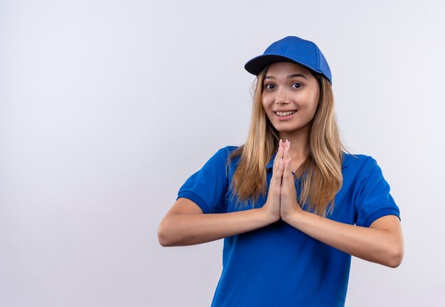 Улыбающаяся молодая доставщица в синей форме и кепке, показывающая жест молитвы, изолирована на белой стене с копией пространства