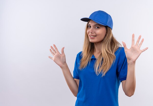 Smiling young delivery girl wearing blue uniform and cap spreads hands isolated on white wall with copy space