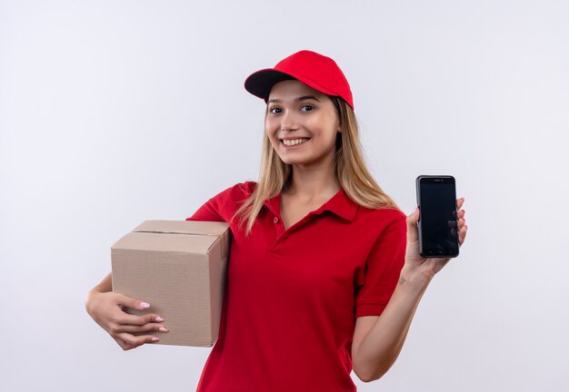 Smiling young delivery girl wearing red uniform and cap holding box and phone isolated on white