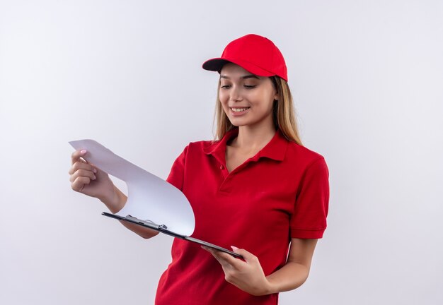 Smiling young delivery girl wearing red uniform and cap holding and flipping through clipboard isolated on white