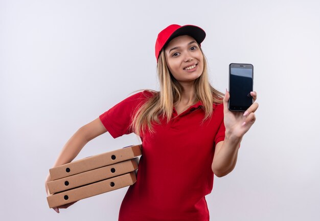 赤い制服と帽子を身に着けている笑顔の若い配達の女の子-白で隔離の電話とピザの箱を保持します。