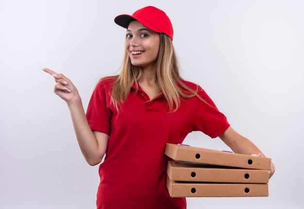 La giovane ragazza sorridente di consegna che porta l'uniforme rossa e la scatola della pizza della tenuta del cappuccio e indica al lato isolato sulla parete bianca