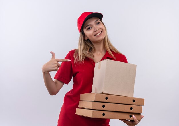 Smiling young delivery girl wearing red uniform and cap holding and points to many boxes isolated on white