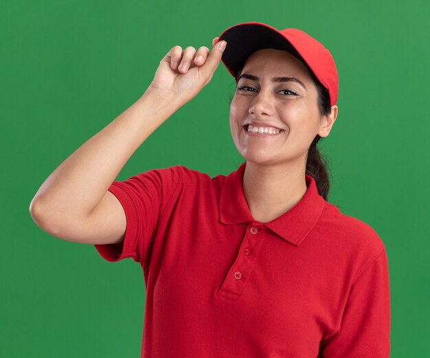 Улыбающаяся молодая доставщица в униформе и кепке, держащая кепку, изолированную на зеленой стене