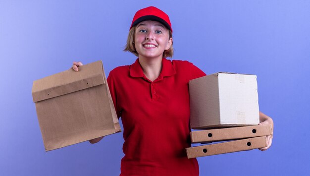 Smiling young delivery girl wearing uniform and cap holding paper food bag with pizza boxes isolated on blue wall