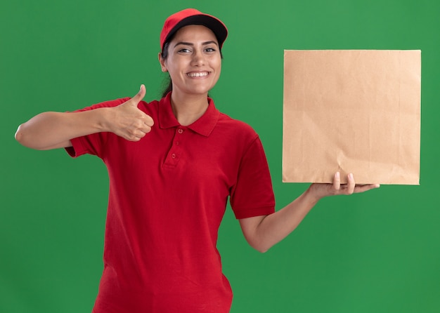緑の壁に分離された親指を示す紙の食品パッケージを保持している制服と帽子を着て笑顔の若い配達の女の子