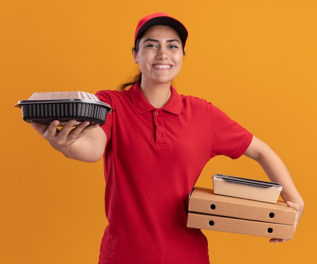Smiling young delivery girl wearing uniform and cap holding pizza boxes and holding out food containers at front isolated on orange wall