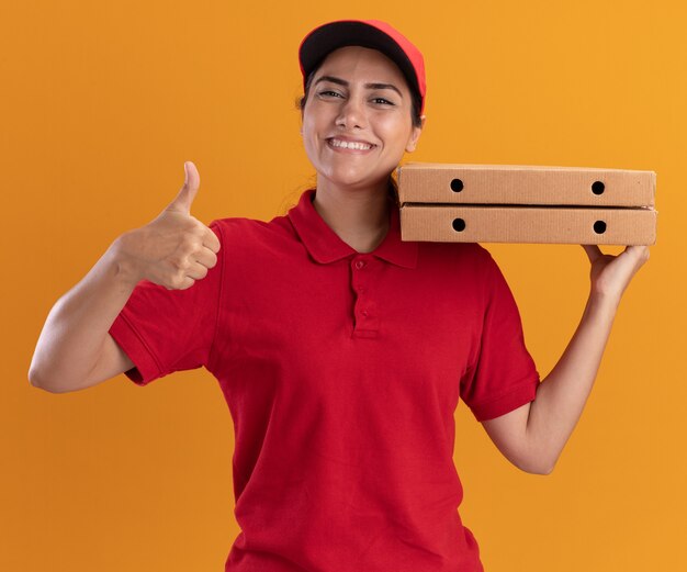 Улыбающаяся молодая доставщица в униформе и кепке держит коробки с пиццей на плече, показывая большой палец вверх изолированно на оранжевой стене