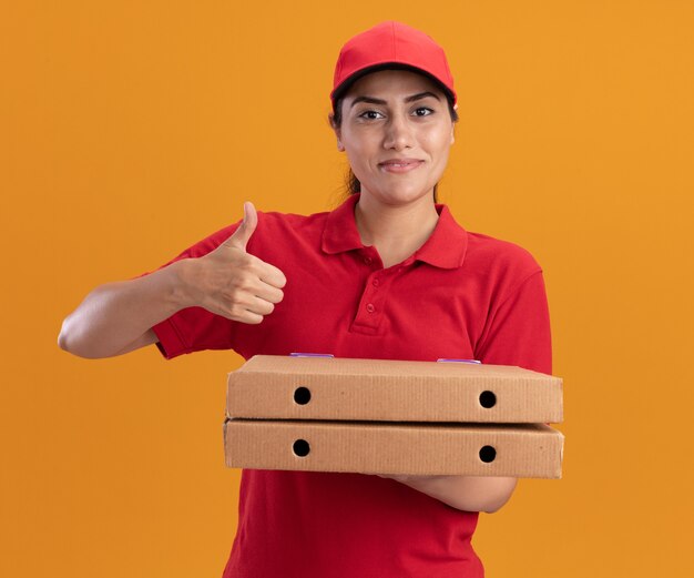 オレンジ色の壁に分離された親指を示すピザの箱を保持している制服と帽子を身に着けている若い配達の女の子の笑顔