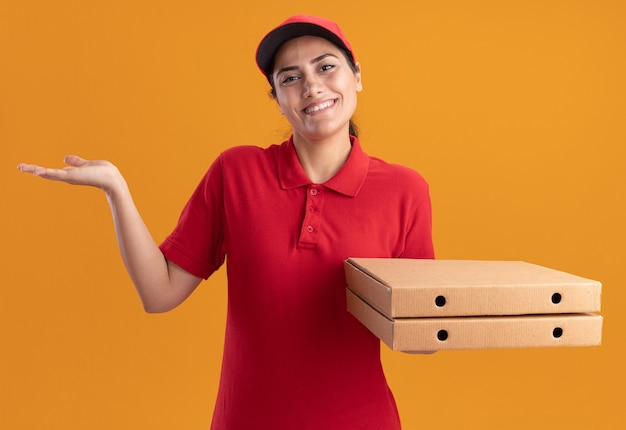 Sorridente giovane ragazza di consegna indossando l'uniforme e berretto tenendo le scatole per pizza diffusione mano isolata sulla parete arancione