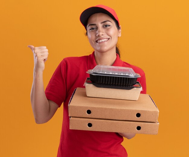 Улыбающаяся молодая доставщица в униформе и кепке, держащая коробки для пиццы с пищевыми контейнерами, указывает сзади, изолированную на оранжевой стене с копией пространства