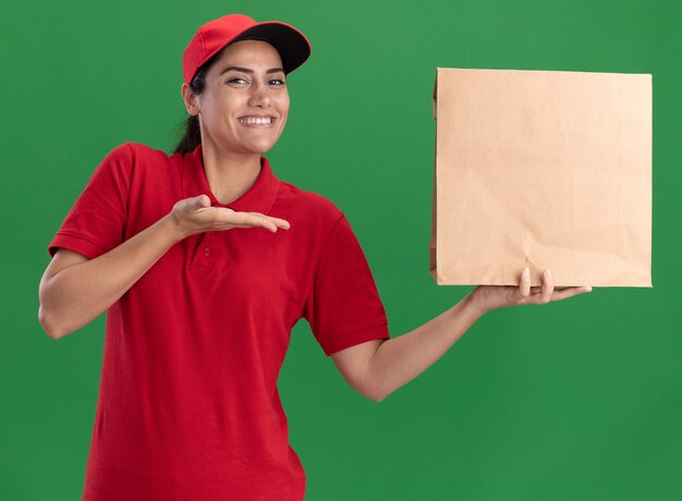 Smiling young delivery girl wearing uniform and cap holding and points at paper food package isolated on green wall