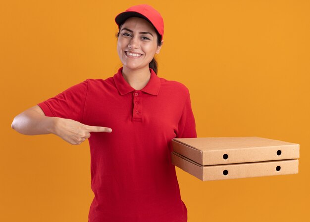 Smiling young delivery girl wearing uniform and cap holding and points at pizza boxes isolated on orange wall