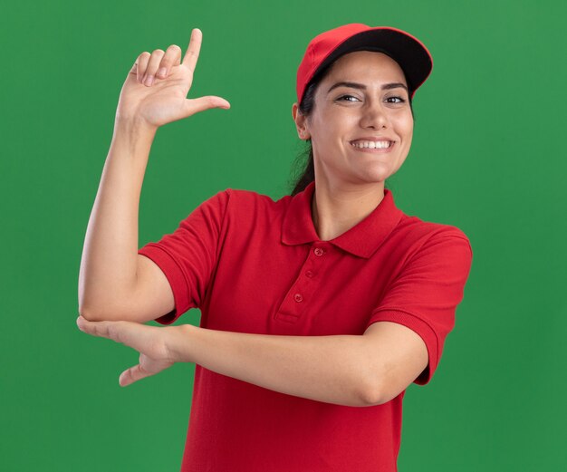 Smiling young delivery girl wearing uniform and cap showing pistol gesture isolated on green wall